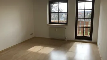 Leeres Zimmer mit Laminatboden, großem Fenster und Zugang zu einem französischen Balkon.