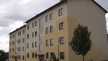 Mehrfamilienhaus mit beiger und gelber Fassade unter bewölktem Himmel und einem Baum davor.