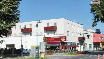 Modernes Mehrfamilienhaus mit Geschäften im Erdgeschoss und roten Balkonen an einer belebten Straße.