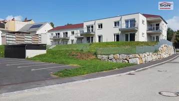 Modernes Mehrfamilienhaus mit Balkonen, Parkplätzen und einer Natursteinmauer in sonniger Lage.