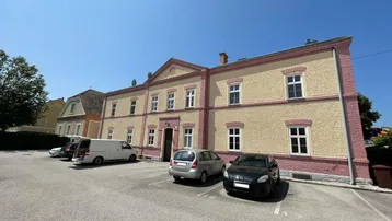 Mehrfamilienhaus mit traditioneller Architektur, Parkplätzen vor der Fassade und Bäumen unter blauem Himmel.