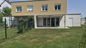 Modernes Reihenhaus mit Garten, Terrasse und Vordach unter blauem Himmel.