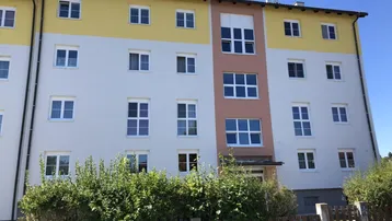 Mehrfamilienhaus mit farbiger Fassade, vielen Fenstern und gepflegter Hecke im Vordergrund.