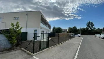 Modernes Mehrfamilienhaus mit Parkplätzen und eingezäuntem Grundstück unter bewölktem Himmel.
