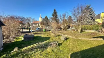Weitläufiger Gartenbereich mit Blick auf einen Kirchturm in der Ferne unter blauem Himmel.