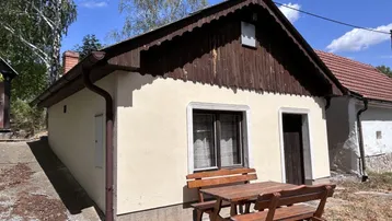 Kleines Landhaus mit Holztisch und Bänken im Vorgarten unter blauem Himmel.