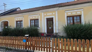 Gelbe Fassade eines traditionellen Hauses mit Holzzaun und grünen Büschen im Vorgarten.