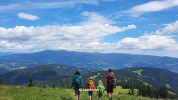Familie wandert auf einem Bergkamm mit weitem Blick über die umliegende Berglandschaft unter blauem Himmel.