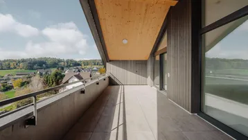 Langer Balkon mit Fliesenboden und Holzdach, bietet weite Aussicht auf die grüne Umgebung.