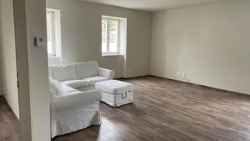 Helles Wohnzimmer mit Holzboden, weißen Wänden und zwei Fenstern, teilweise möbliert.