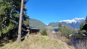 Rustikales Holzhaus in alpiner Landschaft mit schneebedeckten Bergen im Hintergrund unter blauem Himmel.