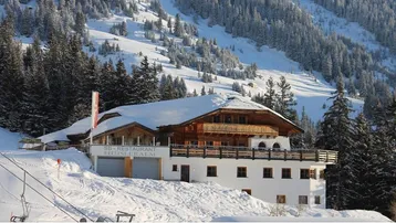 Schneebedecktes Berghaus mit traditioneller Holzarchitektur, umgeben von verschneiten Tannenwäldern und Skipisten.
