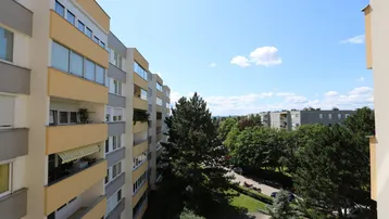 Blick von einem Balkon auf umliegende Wohngebäude, Bäume und die Straße unter blauem Himmel.