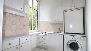 Kleine Küche mit weißen Schränken, Fliesenboden und Fenster, ausgestattet mit Waschmaschine und Kühlschrank.