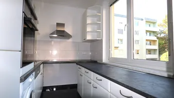 Funktionale Küche mit weißen Schränken, dunklen Arbeitsplatten und Blick auf das Gebäude.