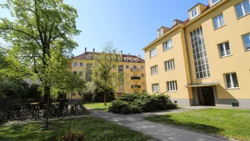 Gelbes Mehrfamilienhaus mit grüner Gartenanlage und Fahrradabstellplätzen unter blauem Himmel.