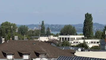 Blick über Dächer mit Solaranlagen auf einen See und bewaldete Hügel in der Ferne.