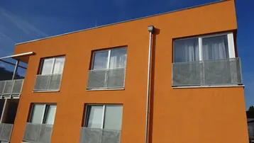 Moderne Gebäudefassade in leuchtendem Orange mit großen Fenstern und französischen Balkonen unter blauem Himmel.