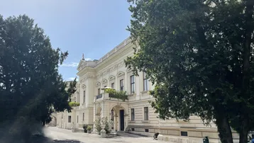 Prächtiges historisches Gebäude mit klassischer Architektur, umgeben von altem Baumbestand und einem gepflasterten Vorplatz.