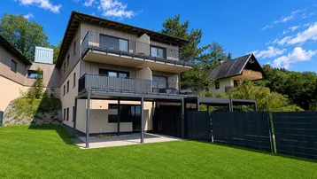 Modernes Mehrfamilienhaus mit Holzfassade, Balkonen und gepflegtem Rasen in bergiger Umgebung.