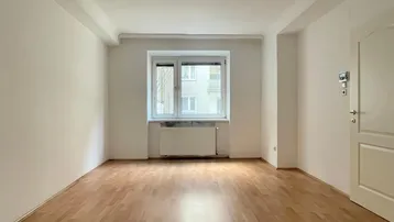 Helles Zimmer mit Parkettboden, weißen Wänden und einem Fenster, das viel Tageslicht hereinlässt.