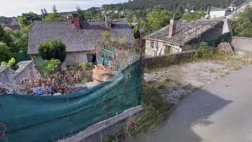 Blick auf zwei ältere, baufällige Häuser mit verwildertem Garten und teilweisem Dacheinsturz, umgeben von Hügellandschaft.