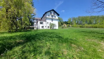Weitläufige Grünfläche vor dem Gebäude mit Waldrand im Hintergrund unter blauem Himmel.