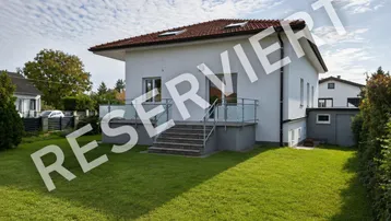 Einfamilienhaus mit weißer Fassade, rotem Ziegeldach und gepflegtem Garten mit Rasenfläche und kleiner Terrasse.