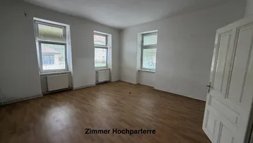 Helles Zimmer im Hochparterre mit Parkettboden, weißen Wänden und mehreren Fenstern.