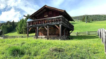 Traditionelle Holzhütte mit Balkon in einer grünen Berglandschaft unter blauem Himmel.