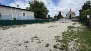 Leeres Kiesgrundstück mit Grasbewuchs, umgeben von grünen Zäunen und Gebäuden im Hintergrund unter bewölktem Himmel.