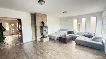 Geräumiges Wohnzimmer mit modernem Holzofen, großem Sofa und Fliesenboden, verbunden mit dem Eingangsbereich.