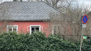 Rotes Haus mit grauem Dach und grüner Hecke an einer Straße.