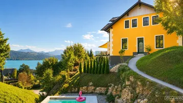 Gelbes Haus mit Pool, gepflegtem Garten und Panoramablick auf See und Berge unter blauem Himmel.