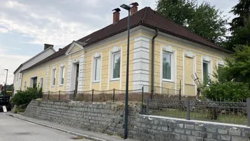 Gelbe Fassade eines älteren Gebäudes mit braunem Dach und Steinmauer im Vordergrund.