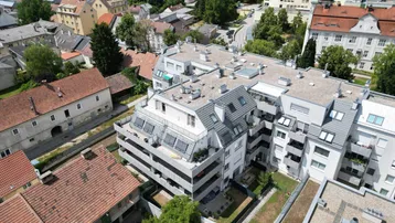 Draufsicht auf das moderne Wohngebäude mit Flachdach und umliegenden Grünflächen sowie älteren Häusern.