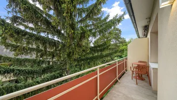 Langer Balkon mit Sitzgelegenheiten und üppigem Grünblick von hohen Bäumen.