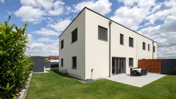 Moderne Hausfassade mit Terrasse und Garten, umgeben von einer Hecke und einem Holzzaun.