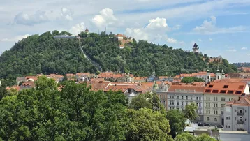 Panoramablick über die Stadt mit dem bewaldeten Schlossberg und historischen Gebäuden unter blauem Himmel.