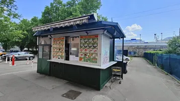 Kleiner Imbissstand mit Markise und Menütafeln, umgeben von einem Parkplatz und Bäumen unter blauem Himmel.