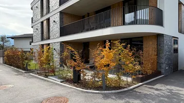 Modernes Mehrfamilienhaus mit Stein- und Holzfassade sowie Balkonen und begrüntem Vorgartenbereich.