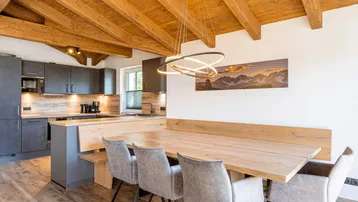 Moderne offene Küche mit dunklen Schränken, Holzelementen und einem großen Esstisch unter Holzbalkendecke.