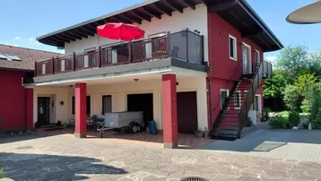 Rotes Haus mit Balkon, Außentreppe und gepflastertem Vorplatz unter blauem Himmel.