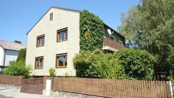 Vorderansicht des Hauses mit heller Fassade, Holzzaun und üppiger Begrünung unter blauem Himmel.