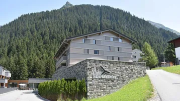 Modernes Gebäude mit Stein- und Holzfassade, umgeben von grünen Bergen und Wäldern.