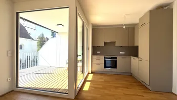 Heller offener Wohnbereich mit moderner Einbauküche, Holzboden und Zugang zu einer sonnigen Terrasse.