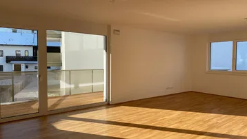 Helles, leeres Wohnzimmer mit Parkettboden, großen Fenstern und Zugang zum Balkon.