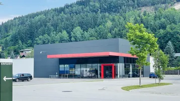 Modernes Geschäftsgebäude mit großer Glasfront und rotem Vordach vor bewaldetem Hügel und Parkplatz.