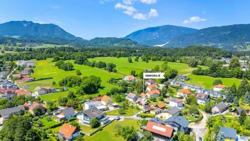 Luftaufnahme einer Wohnsiedlung mit grünen Feldern und Bergen im Hintergrund, die Immobilie ist markiert.