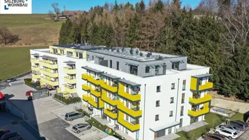 Modernes Wohngebäude mit gelben Balkonen, umgeben von Grünflächen und Bäumen, mit Photovoltaikanlage auf dem Dach.